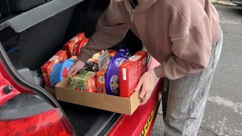 Elder Tree Befriending A car boot is being filled with hampers made from cardboard. The items include tea, crackers, chocolate and biscuits. The car is red and a young woman is lifting the hamper into the car.