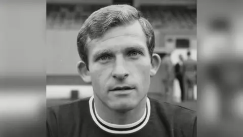 A black and white image of Tony Bentley, who has light hair and is looking at the camera expressionless. The picture has been taken in front of a stand at the stadium.