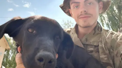 Tindal-Draper family handout Callum Tindal-Draper wearing military camouflage clothing while holding a black dog.