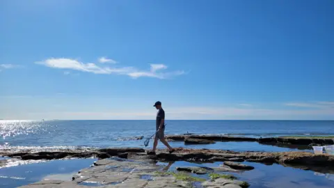 Maria A man wearing shorts, tshirts and a cap walks across rocks carrying a small net. The rocks are on the edge of the sea beyond that is bright blue in the sun. He is wearing black shoes and there are several rock pools. The sky is blue with one fluffy white cloud. 