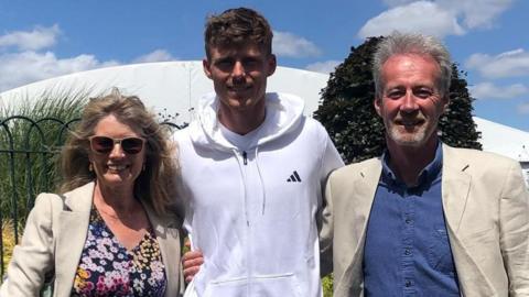 Ian and Jan Watson stand either side of Billy Harris. They have their arms around each other. Billy is wearing a white zipped jumper and Jan and Ian are wearing cream blazers.