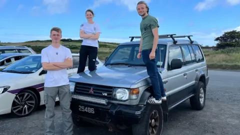 Fergus standing beside the car and Daisy and Emile standing on the wheels