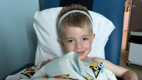 A young boy with blonde hair wearing a white bandage around his head. He is smiling at the camera while sitting on a hospital chair. 