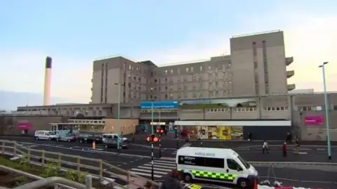 BBC A picture of Derriford Hospital from the car park. There is an ambulance parked on the road. There is a zebra crossing on the road. At the front of the hospital is a Marks & Spencer food hall.