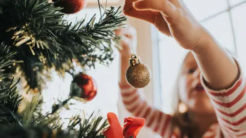 A girl is wearing a red and white stripy long top and is carefully putting a sparkly gold bauble on a Christmas tree.