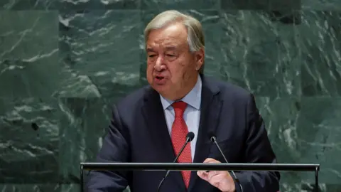 UN Secretary General Antonio Guterres giving a speech at the United Nations in New York. He is standing against the distinctive green marbled backdrop at a lectern, wearing a red tie, dark suit jacket and blue shirt