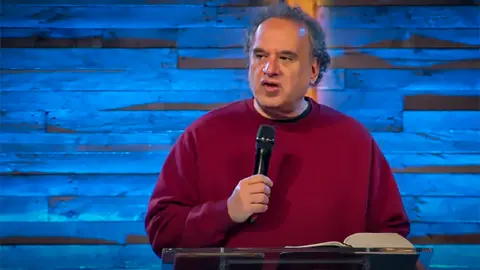 Mike Pilavachi wearing a red jumper and holding a microphone. In front of him is a lectern with an open book on it and behind him is a blue backdrop.