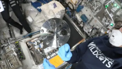 A circular metal part spins in zero gravity as two astronauts look on wearing masks and goggles