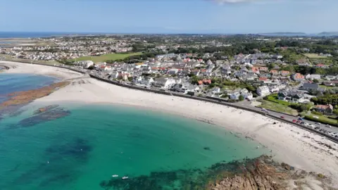 A drone shot of Guernsey. The sea is a light blue colour with white sands near to it.