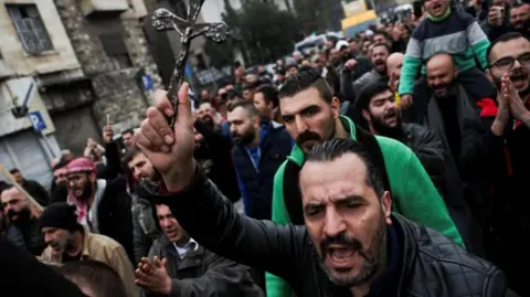 Reuters A shot of a crowd of protesters marching through a street. In the foreground, a bearded man holds up a small cross as he yells.