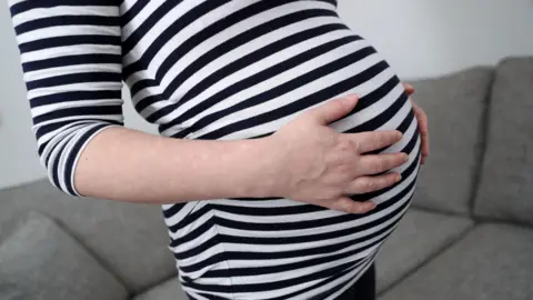 A heavily pregnant woman wearing a black and white long sleeve t-shirt holds her stomach