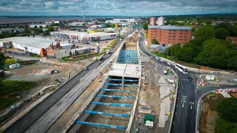 Aerial view of the road work showing an underpass being excavated with blue girders all the way along and a bridge in the middle