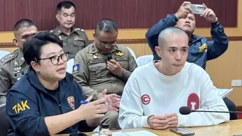 Wang Xing with a shaved head sitting at a table. Beside and around him are Thai officials in uniform.