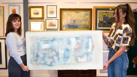 Holly Hammond is standing to the left and holding the picture by Patrick Heron, with Coralie Thomsom holding it on the right. In the background are other pictures from artists mounted on the wall 