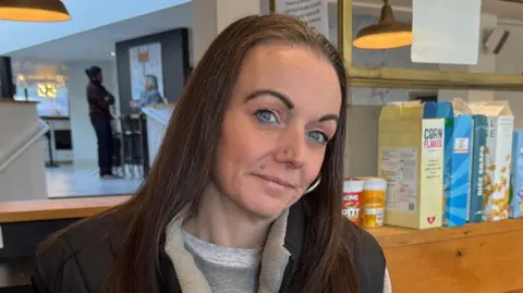 Kelly is in the bar area of a hotel which is being used as a kitchen. She is smiling at the camera and holding a mug. She is wearing a blue body warmer over a grey sweatshirt. There is a row of cereals behind her