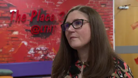 Janine Craven, a woman with dark long hair and black rimmed spectacles, looks away from the camera. She is in a room with a large red wall and blue sofa behind her.