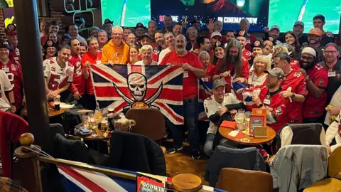 About 50 Buccaneers fans in a bar in London at one of their watch parties. All are crowded together and wearing the team's jersey. Some are holding Great Britain flags, which also have the Buccaneers logo in the middle. 