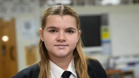 John Fairhall/BBC Scarlett, a Year 8 pupil. She has long fair hair, pulled back from her head and falling over either shoulder. She is wearing a dark jacket, dark tie and a white shirt. She is sitting in a classroom, blurred out behind her