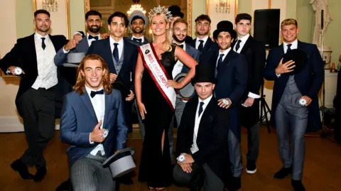 @mrenglanduk Bal poses for a picture with the other finalists, all in top hat and tails. Miss England is in the centre, wearing her crown, sash, and long black dress.