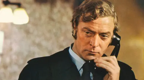 A still from the film Get Carter, showing Michael Caine as Jack Carter. He is holding a black telephone receiver to his ear and wearing a light blue shirt, a black tie and a black raincoat