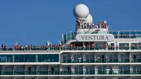 Getty Images Close-up of passengers standing on the top deck of a cruise ship. A large name plate reading 'Ventura' is on the side of the ship. Cabin balconies can also be seen.