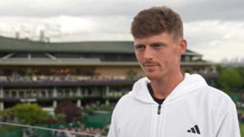 Billy Harris wears a white zip-up jumper, he stands on a balcony at Wimbledon. 