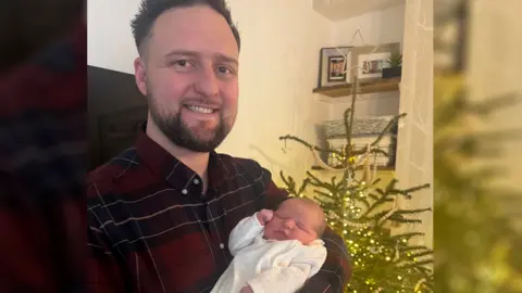 A tiny infant is being held by her dad in front of a Christmas tree. he is wearnign a check shirt and she is in a white knitted baby jumper