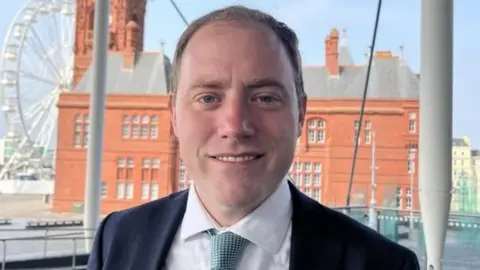 BBC A head and shoulders picture of Rhys ab Owen, wearing a suit, shirt and tie, stood with Cardiff Bay's terracotta-glazed Pierhead building.