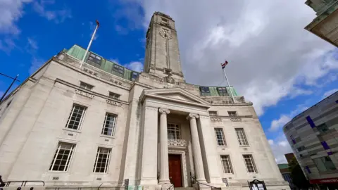 A picture of Luton Council Headquarters in Luton. It's a grey-ish white building. It has a neoclassical style with art deco detailing and the sky is blue behind it aside from one greyish cloud.