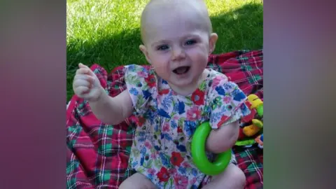 Family Zoey pictured when she was a baby garden sat on a red and green checked picnic blanket.

She has a blue and pink floral dress on and has a green hoop tucked under her left armpit