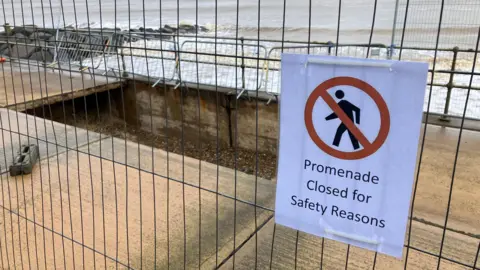 Metal fencing surrounds a large, rectangular-shaped hole in a concrete promenade with a sign on the fence reading "promenade closed for safety reasons". Waves and a rock groyne can also be seen in the background. 