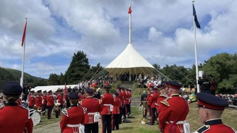 Members of a military band wearing red jackets with gold trim and black trousers and caps are lined up with their backs to the camera in front of Tynwald Hill. The grass-covered circular hill is tiered with stone steps leading up to it, with people sitting on chairs on each tier. It is topped by a white canopy and a flagpole flying a red Manx flag and, the hill is also flanked by to other flagpoles flying Manx and Tynwald flags.