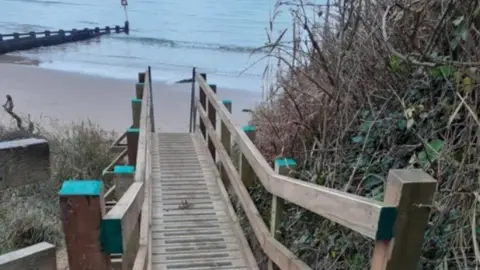 Swanage Town Council Wooden boardwalk with handrails, shrubs either side and beach, sea and groyne seen off the end. 