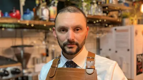 A man with a short beard, wearing a brown leather apron over a white shirt standing in a coffee shop. The background is out of focus but shelves and equipment can be made out