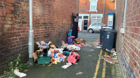 An alleyway leading to a residential street with terraced houses. A number of items have been dumped on the left of the alleyway. There are two black bins next to the wall on the right, opposite an open black gate. A silver car can be seen in the background parked next to the terraced houses.