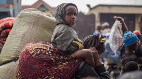 A young boy sitting on an adult's shoulders stares at the camera as residents carry their belongings as they flee from Kibati, where fighting has intensified, towards the city of Goma on January 26, 2025.