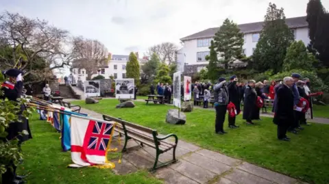 People stood in a park during a military memorial service. A group of people on the left-hand side are holding flags on poles which have been lowered to the ground. Other people stood on a patch of grass have poppy wreaths in their hands.
