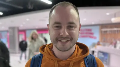 Craig Ballentine at Dublin Airport, he has short light hair and a beard. He is smiling at the camera and wearing an orange hoodie and a blue back pack 