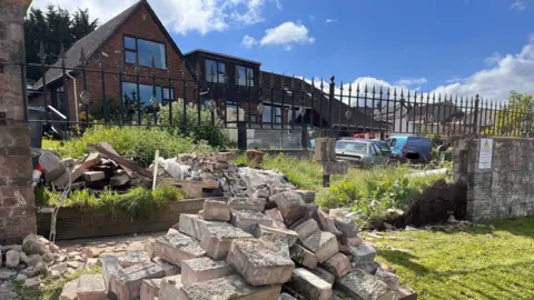 A pile of bricks and other debris in front of a black metal fence with a house in the background