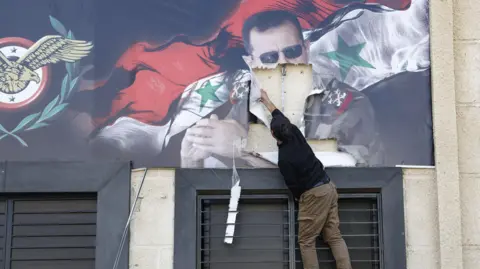 EPA A man tears down a banner showing Bashar al-Assad and the Syrian flag at Mezzeh airbase, on the outskirts of Damascus (16 December 2024)