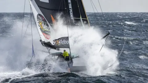 Sam Goodchild Racing A sailing boat in rough seas with the word Sodebo on a sail and a sailor in a yellow top in the cockpit.