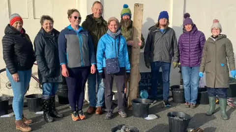 Volunteers pictured wearing warm winter clothing outside. There are black buckets around them with litter inside. 