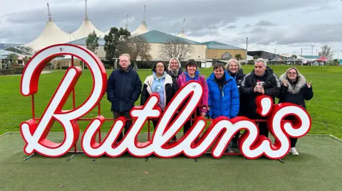 My Day Care Services Eight people standing behind a large Butlin's sign in front of a field and the holiday park's buildings behind