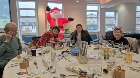 A table laid out with Christmas Dinner decorations such as crackers, and guests sitting around the table.