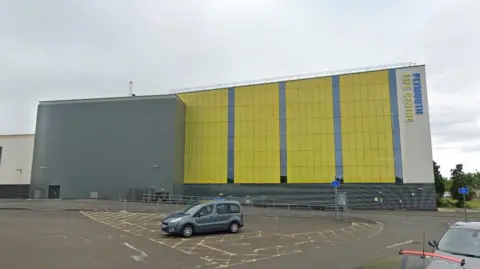 Plymouth Life Centre building exterior. The building is green and grey. A car park is in front of the building. A grey car is parked in a disabled bay.