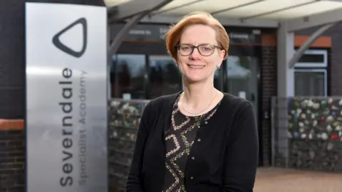 A woman with short red hair and glasses. She is smiling and wearing a black cardigan on top of a black patterned shirt.