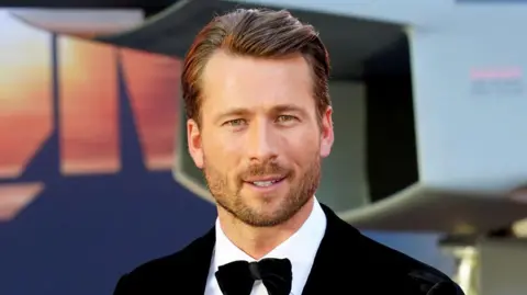 Glen Powell, dressed in a suit with bow tie, on the red carpet for a film premiere. He has dark brown hair and is unshaven with stubble.