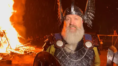 Calum Grains wears a winged viking helmet and silver armour. He holds a shield in his right hand. He stands in front of a burning long ship. Flames lick high up into the sky and embers rain down behind him.