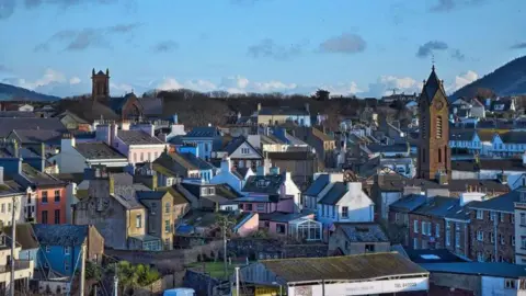 A high up view of rooftops with two church towers and a hill in the background with a clear blue sky.