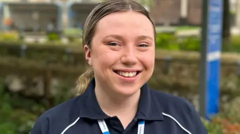 Cambridge University Hospitals NHS Foundation Trust Sian Leader wearing a dark blue medical uniform. Her mid-brown hair is pulled back from her forehead and tied into a pony tail. She is smiling at the camera. 